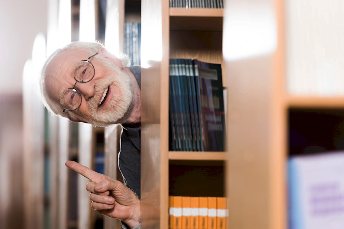 Starszy mężczyzna z brodą i okularami wygląda zza regału z książkami w bibliotece, wskazując palcem.